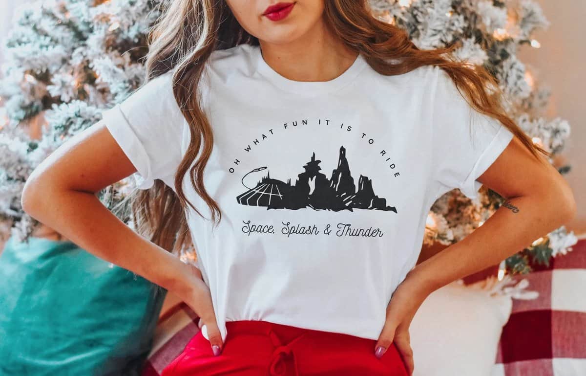 In front of a sparkling Christmas tree, a Disney fan shows off the ultimate festive park style with a white shirt featuring silhouettes of iconic Walt Disney World attractions and themed text—perfect inspiration for holiday adventures in the parks.
