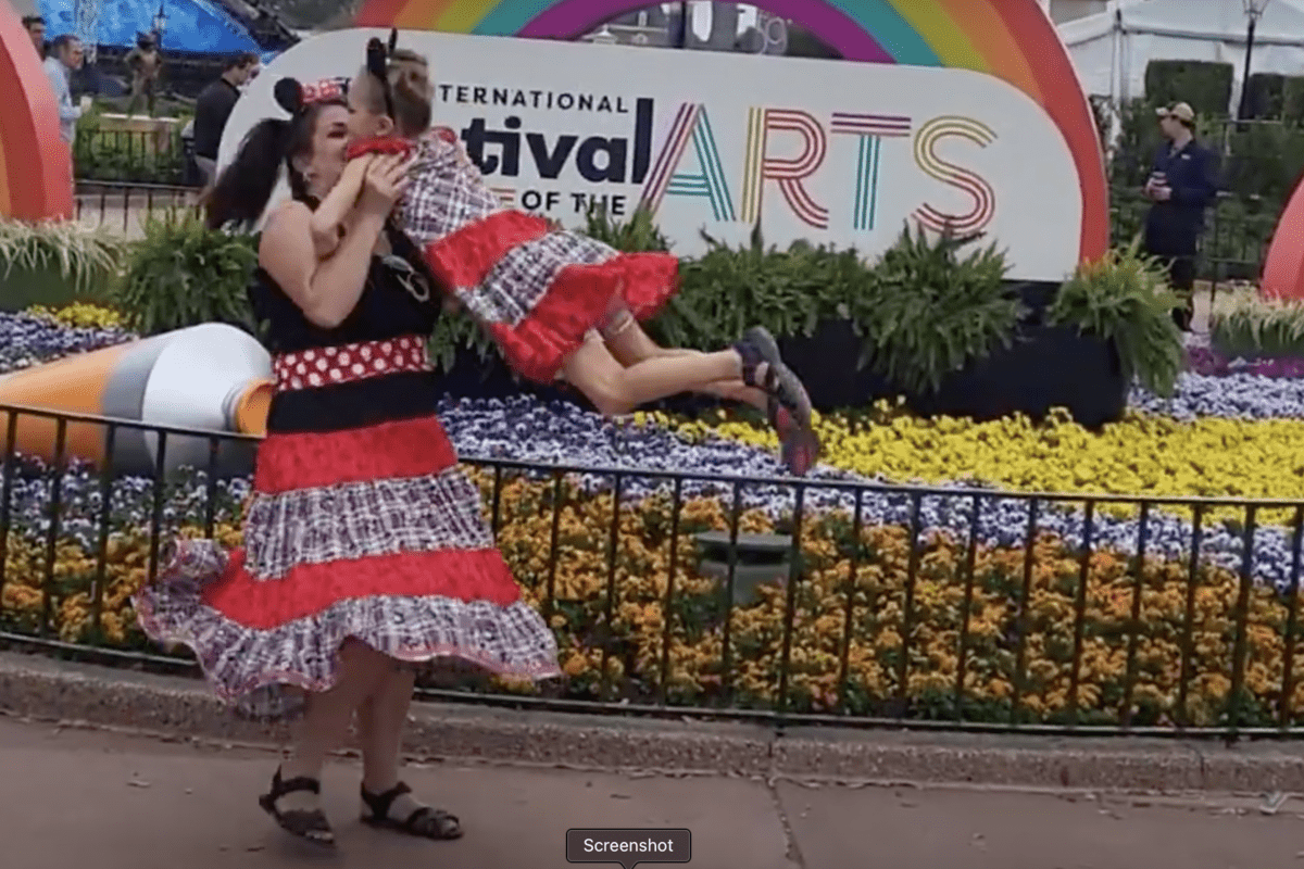 A Disney World guest in a vibrant red dress twirls her child with joy in front of the eye-catching Festival of the Arts sign and a lush flower bed, highlighting practical yet stylish park attire amidst the festivities.