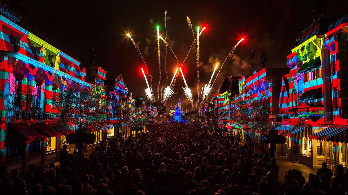 Main Street, U.S.A. packed with excited guests under the glow of vibrant building lights, as dazzling fireworks explode above Cinderella Castle in the distance while Disney’s soundtrack accidentally plays music from a different fireworks show.