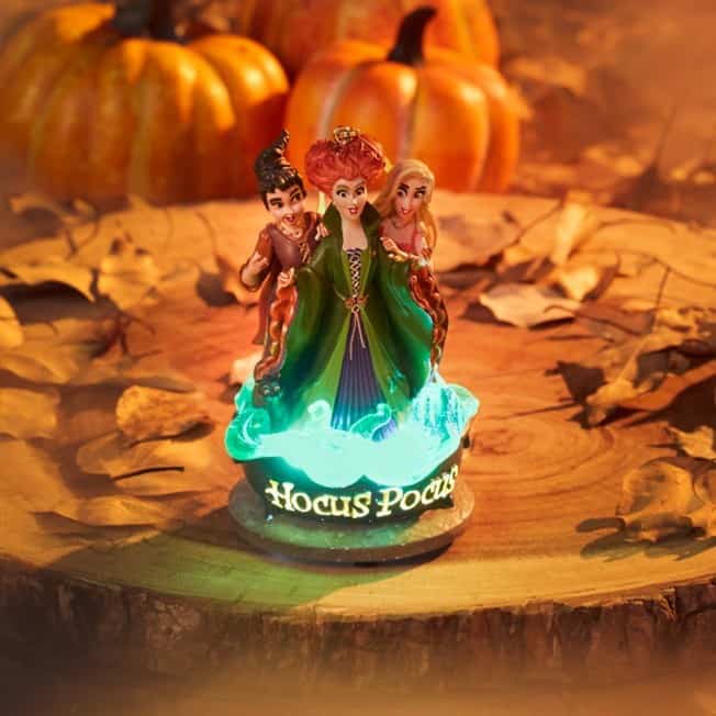 The Sanderson Sisters from “Hocus Pocus” gather atop a glowing decorative base, surrounded by pumpkins and colorful autumn leaves, capturing the magic of the Halloween season for fans of Disney’s iconic film.