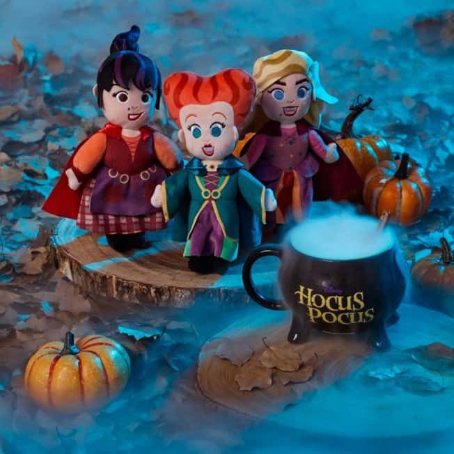 The Sanderson Sisters plush dolls from Hocus Pocus are displayed beside a bubbling cauldron mug and festive mini pumpkins on a bed of autumn leaves, evoking the magic of the film—perfect for enhancing your Hocus Pocus collection at Disney Parks or themed events.