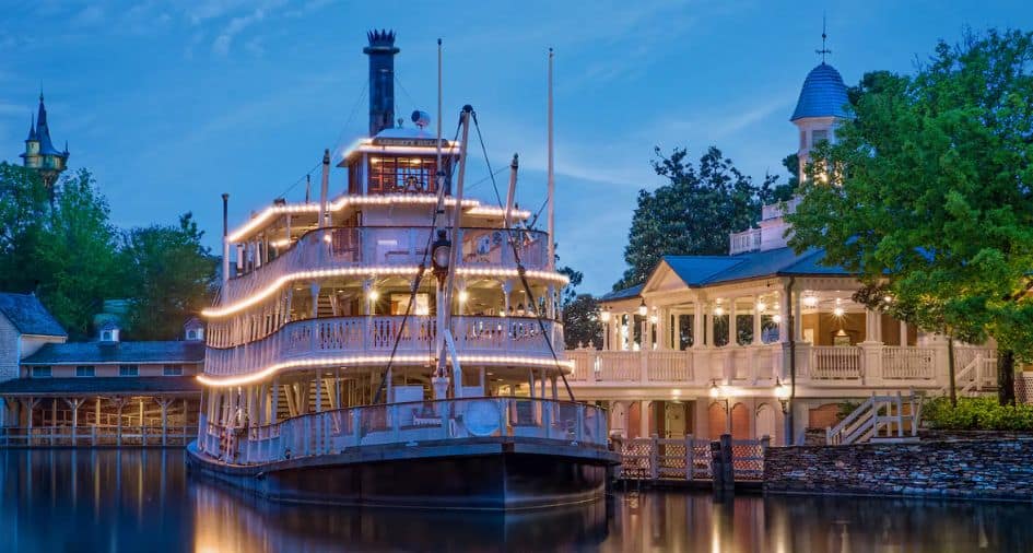 The Liberty Belle riverboat, illuminated with twinkling lights, is docked beside the riverside building at dusk in Liberty Square. Its reflection shimmers on the calm water, setting a peaceful scene as Magic Kingdom winds down for the night.