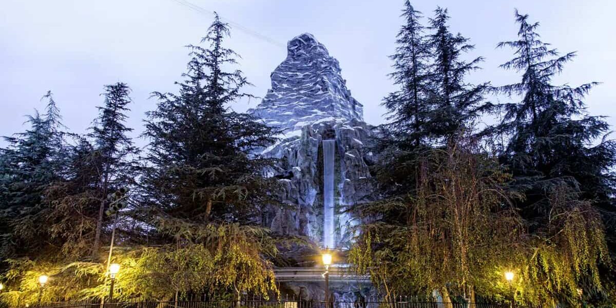 The iconic Matterhorn mountain towers above with a snowy peak and cascading waterfall, framed by majestic evergreens and twinkling street lamps as evening falls—a serene scene that Disneyland fans now glimpse while the Matterhorn Bobsleds attraction is temporarily closed.