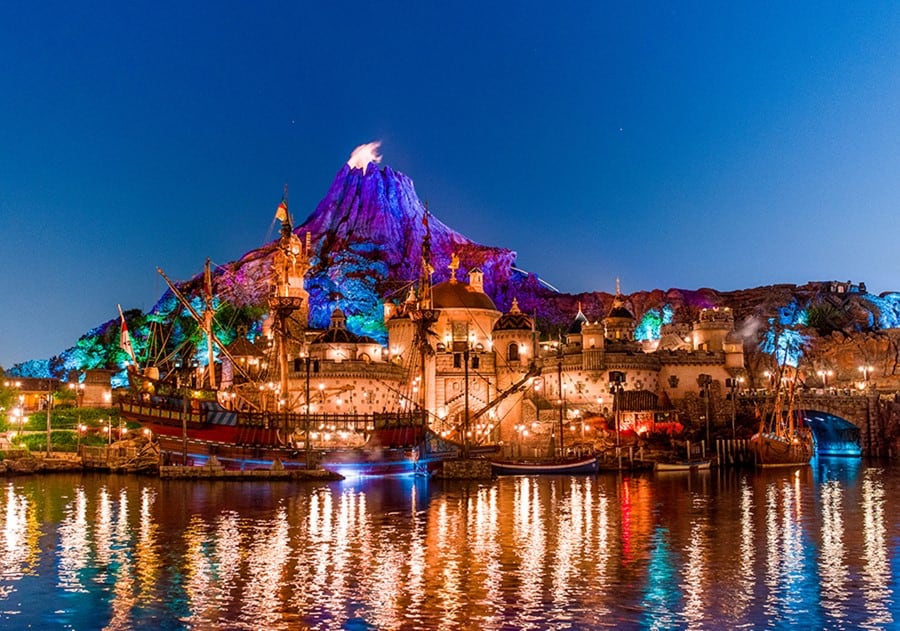 Universal’s iconic Volcano Bay glows at dusk, its towering volcano reflected in peaceful waters, as an excited park visitor playfully joins the fountain display, capturing the magic and energy of the themed paradise.