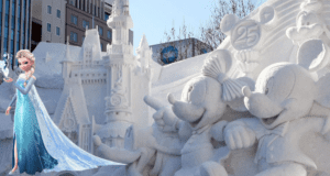 Elsa poses beside intricately sculpted snow figures of Mickey and Minnie Mouse, along with a snowy Disney castle, all set against a city backdrop glowing in sunlight—capturing the enchanting holiday spirit that feels right at home in a Disney theme park’s festive winter celebration.