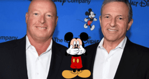 Bob Iger and Bob Chapek, both dressed in suits, smile as they pose with Mickey Mouse—who playfully covers his mouth—in front of a vibrant blue Disney backdrop.