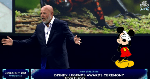 Chapek stands on stage at a Disney event with a dejected Mickey Mouse illustration next to him while disappointed Disney fans in the audience loudly boo.