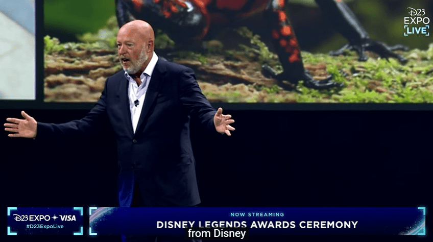 Bob Chapek stands on stage in a suit at the Disney Legends Awards Ceremony during D23 Expo, addressing the crowd as some Disney fans in the audience voice their disapproval by booing.