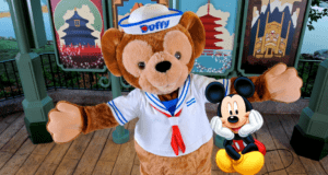 Duffy the Disney Bear, sporting his classic sailor suit, joins Mickey Mouse for a cheerful photo op in front of vibrant, Disney-themed posters at the park.