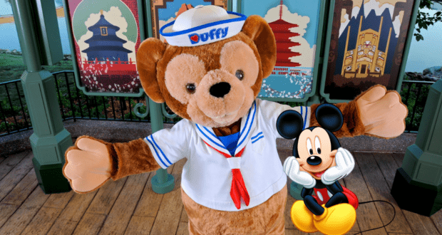 Duffy the Disney Bear, sporting his classic sailor suit, joins Mickey Mouse for a cheerful photo op in front of vibrant, Disney-themed posters at the park.