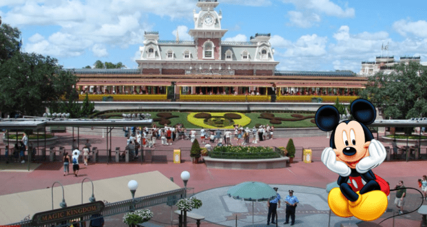 The iconic Magic Kingdom entrance greets guests with a vibrant Mickey Mouse floral display, while a cheerful cartoon Mickey Mouse waves from the bottom right corner, setting the stage for Rope Drop excitement as fans gather to kick off their day of Disney magic.