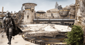 The Mandalorian poses with Grogu in front of the Millennium Falcon at Star Wars: Galaxy’s Edge, surrounded by the outpost’s iconic rocky structures, creating a must-see photo op for fans visiting the immersive themed land.