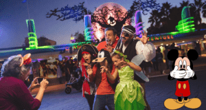 Families dressed in creative costumes gather at the Disneyland entrance at dusk, smiling for photos as a playful Mickey Mouse graphic appears in the corner, ready to celebrate Oogie Boogie Bash with their tickets in hand.