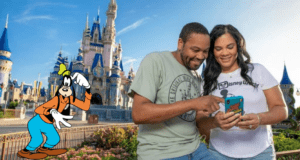 A smiling couple checks their phone in front of Cinderella Castle as Goofy waves nearby, capturing the excitement of planning a Disney Parks vacation.