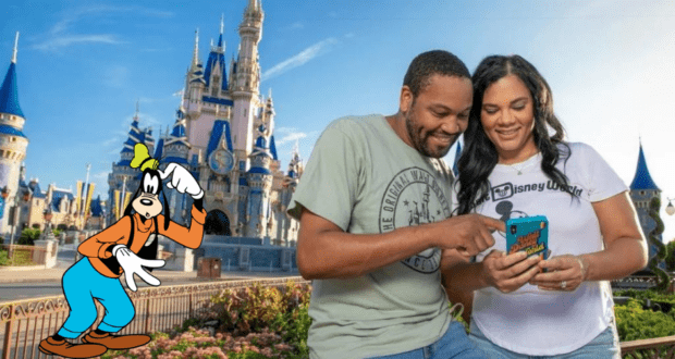 A smiling couple checks their phone in front of Cinderella Castle as Goofy waves nearby, capturing the excitement of planning a Disney Parks vacation.