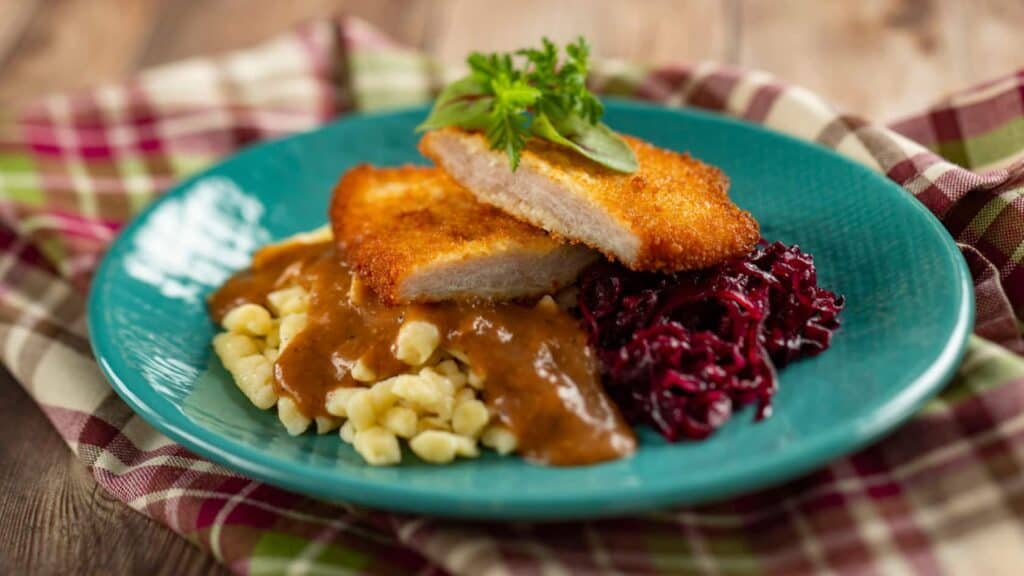 Savor a breaded pork cutlet served atop noodles, draped in rich brown gravy and paired with vibrant red cabbage and fresh herbs, beautifully presented on a blue plate—capturing the festive EPCOT International Festival of the Holidays experience right at your table.