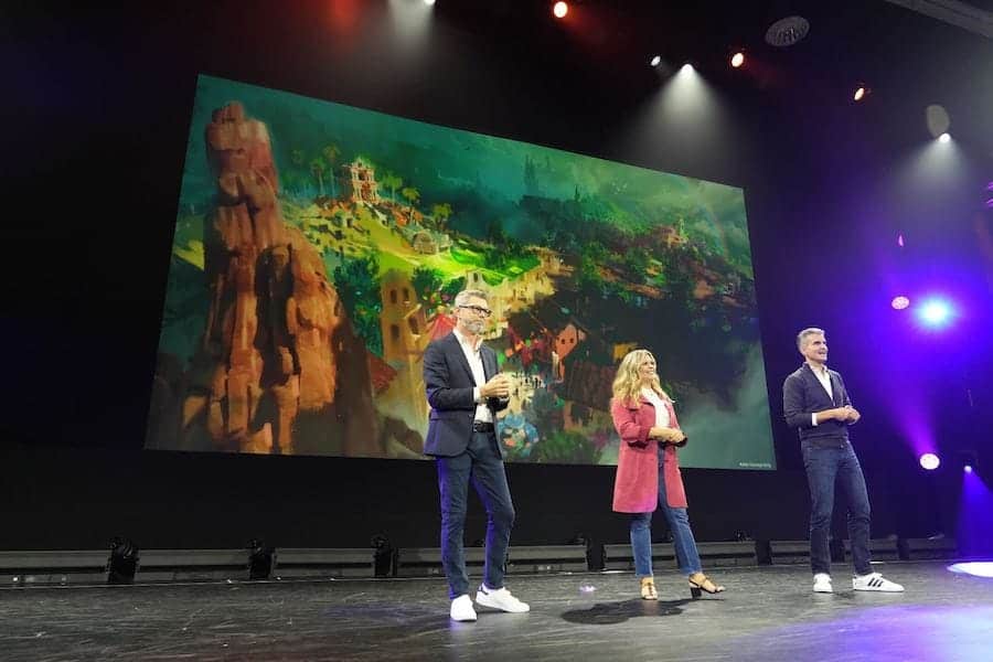 Three presenters stand on a stage with a massive, vividly colored video game world projected behind them—its vibrant look capturing the magical energy and immersive style of an Encanto-themed experience at Magic Kingdom.