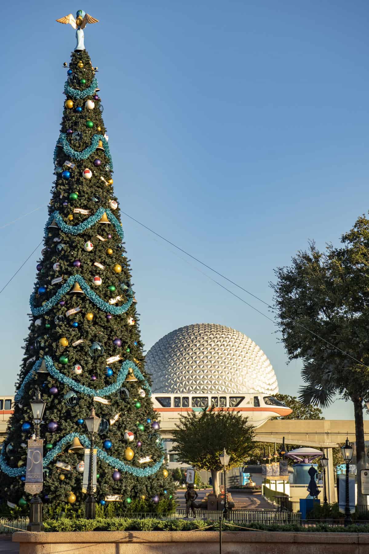 A vibrant Christmas tree gleams in front of Epcot’s iconic Spaceship Earth under bright blue skies, highlighting the cheerful atmosphere of EPCOT Holiday Kitchens for Disney park guests.