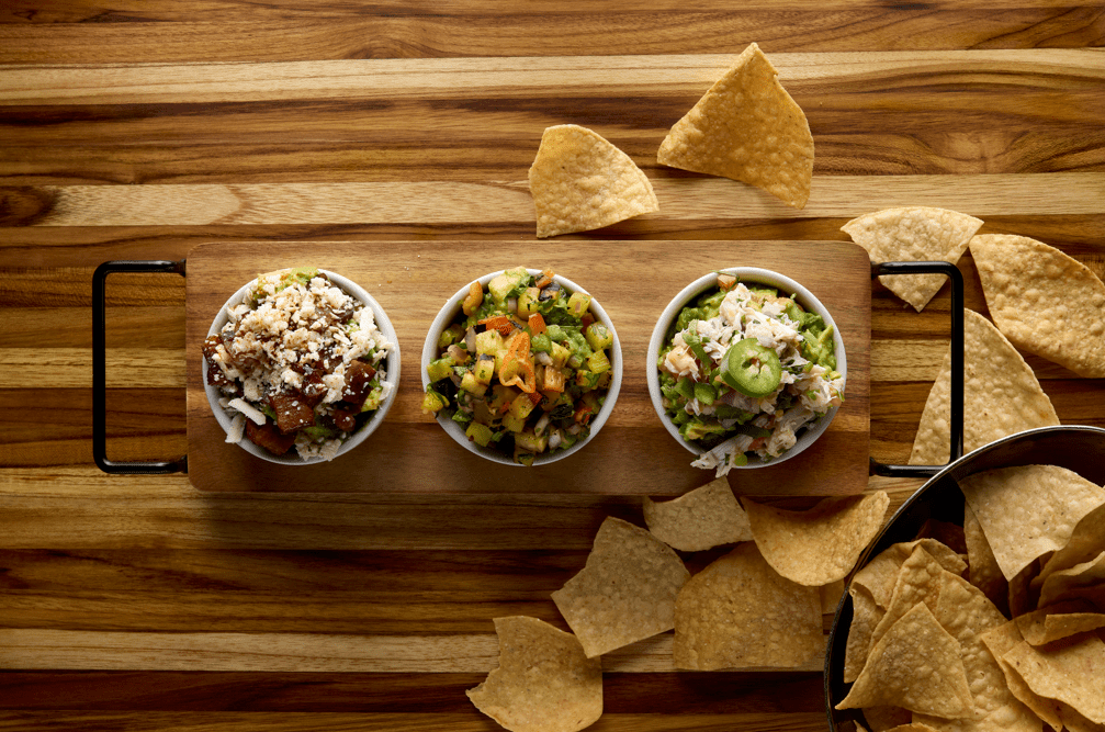 A festive display of three salsa bowls with tortilla chips on a wooden board, surrounded by more chips atop a rustic table—capturing the colorful, lively snack experience found at Rosa Mexicano inside Disney Springs.