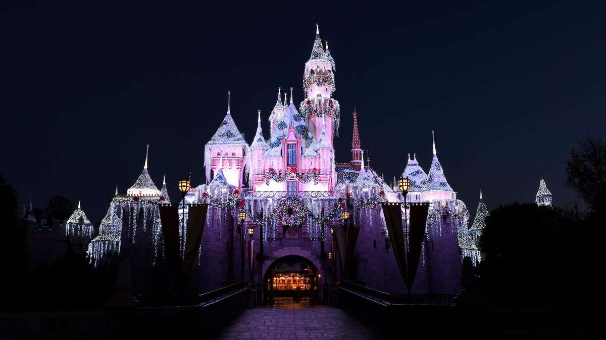 Sleeping Beauty Castle illuminated with vibrant holiday lights and festive decorations stands out against the night sky at Disneyland, setting a magical scene for guests even as attractions like It’s a Small World temporarily close for their popular seasonal layover.
