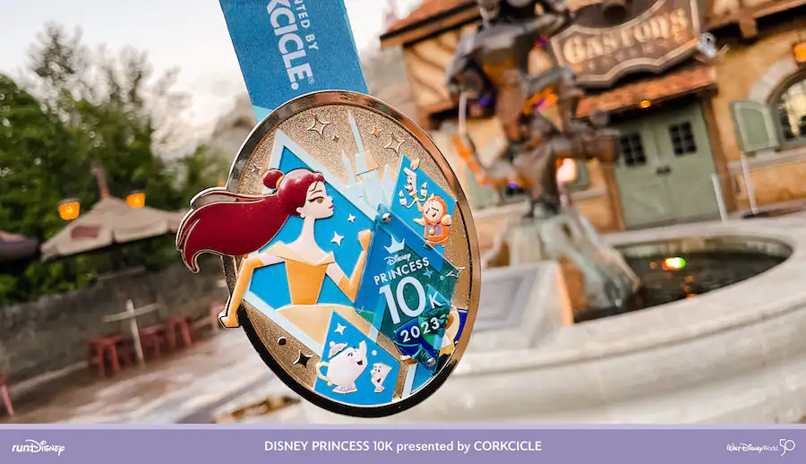 Disney Princess 10K medal featuring Ariel and her undersea friends, displayed in front of a whimsical Disney resort building—a sparkling souvenir from runDisney’s Princess Half Marathon Weekend at Walt Disney World.