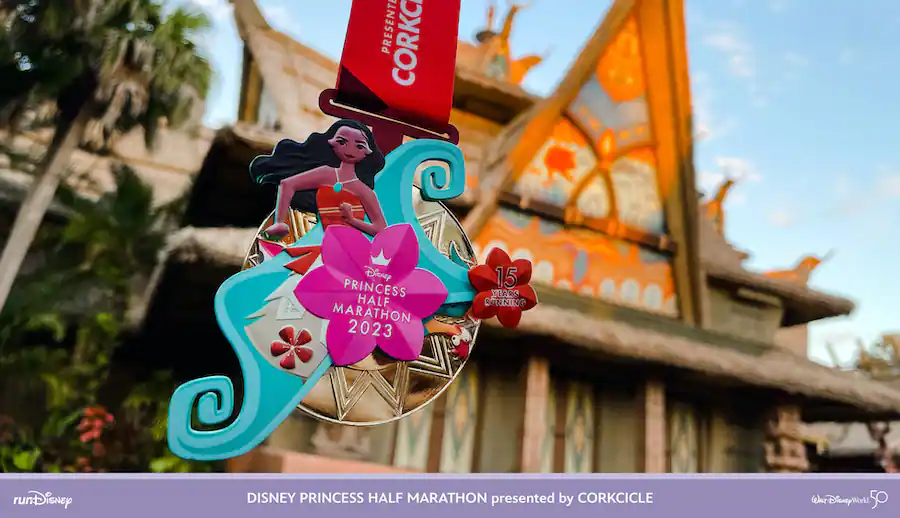 The Disney Princess Half Marathon 2023 medal featuring Moana is proudly displayed in front of a vibrantly themed Disney Parks building, highlighting the excitement and accomplishment of this beloved runDisney event.