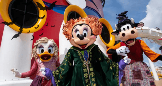 Mickey Mouse, Minnie Mouse, and Goofy show off their Halloween costumes aboard a festively decorated Disney Cruise Line ship, surrounded by colorful décor and flying bats, bringing the spirited magic of Halloween on the High Seas to life.