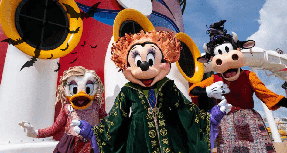Mickey Mouse, Minnie Mouse, and Goofy show off their Halloween costumes aboard a festively decorated Disney Cruise Line ship, surrounded by colorful décor and flying bats, bringing the spirited magic of Halloween on the High Seas to life.