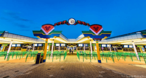 Walt Disney World’s iconic entrance gates illuminated at dusk, with glowing lights and vibrant green queue lines stretching beneath a deep blue sky. Enhanced security presence is noticeable near the entrance after earlier safety concerns were reported.