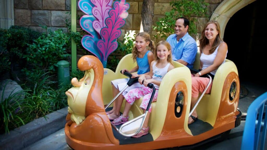 At Disneyland, a family of four beams with delight as they ride the iconic Caterpillar attraction, while nearby, a mom's playful scream adds an extra thrill to the park's lively and magical atmosphere.