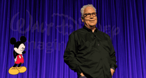 A Disney stage lights up with a vibrant Mickey Mouse illustration as legendary Imagineer Bob Weis is honored, standing beneath the iconic Disney logo on a purple curtain backdrop, celebrating creativity and magic in front of an enthusiastic theme park crowd.