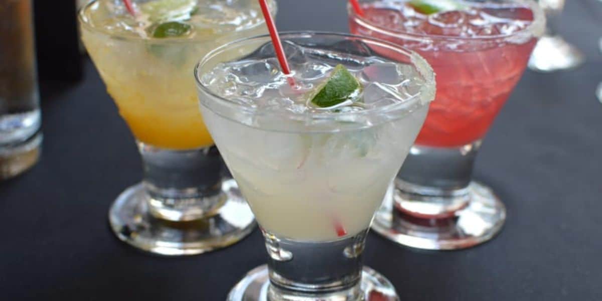 Three cocktails in short glasses, each garnished with a lime wedge and a red straw. The drinks are yellow, clear, and pink, set on a dark table. With the drink prices just as inviting as the vibrant colors, these cocktails are perfect for any occasion.