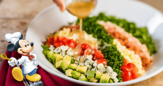Remy from Ratatouille, donning his chef’s hat, stands proudly beside a bowl of freshly chopped salad at Disneyland as tangy dressing is poured over the top, beaming with pride like a star at a Disney Pixar celebration.