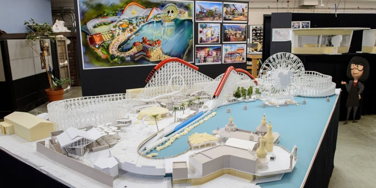 A detailed scale model of an amusement park, inspired by lands in Disney Parks that no longer exist, features roller coasters, a Ferris wheel, and a water area. Behind it are concept art images and a mannequin in glasses and black outfit.