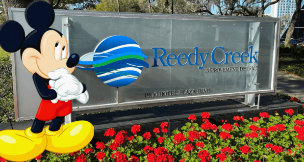 Mickey Mouse greets guests in front of the Reedy Creek Improvement District sign at Disney World, surrounded by vibrant red flowers that capture the playful spirit and magical atmosphere of Reedy Creek.
