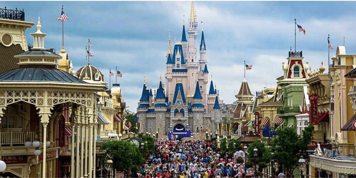 A large crowd gathers on Main Street in front of a blue-and-white fairytale castle at a theme park, surrounded by colorful buildings and American flags—unaware of the hidden utilidors bustling beneath them—under a partly cloudy sky.