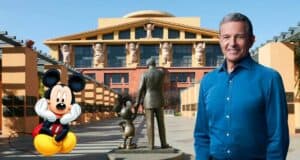 Mickey Mouse poses with a smiling guest beside a decorative statue outside a Disney building adorned with character figures, radiating the energy and excitement of the Bob Iger Town Hall celebration.