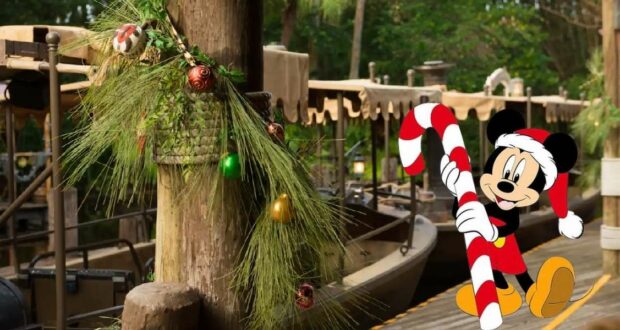 Mickey Mouse sporting a festive Santa hat and holding a candy cane greets guests beside holiday-themed boats adorned with pine garlands and ornaments, capturing the merry atmosphere of Disney’s Jingle Cruise attraction.