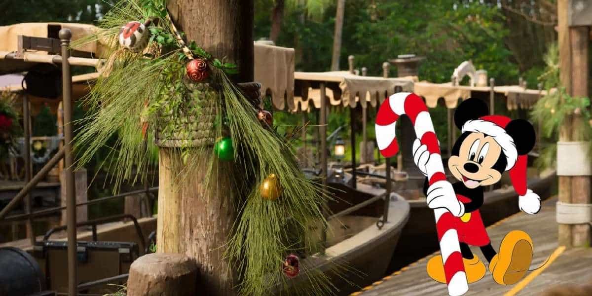 Mickey Mouse sporting a festive Santa hat and holding a candy cane greets guests beside holiday-themed boats adorned with pine garlands and ornaments, capturing the merry atmosphere of Disney’s Jingle Cruise attraction.
