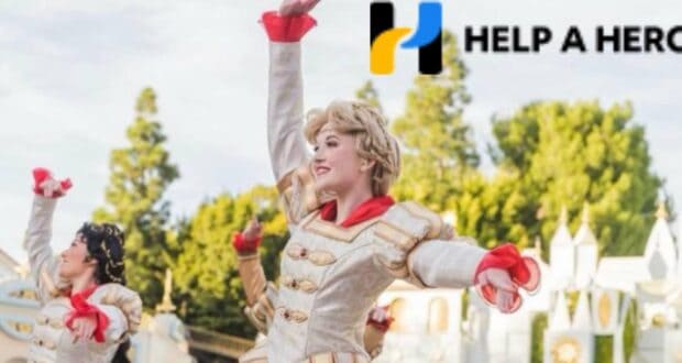 Disney cast members in vibrant, themed costumes perform an energetic dance outdoors as part of a special tribute to late Disney colleagues, with the "Help a Hero" logo displayed in the upper right corner.
