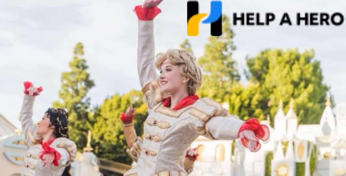 Disney cast members in vibrant, themed costumes perform an energetic dance outdoors as part of a special tribute to late Disney colleagues, with the "Help a Hero" logo displayed in the upper right corner.