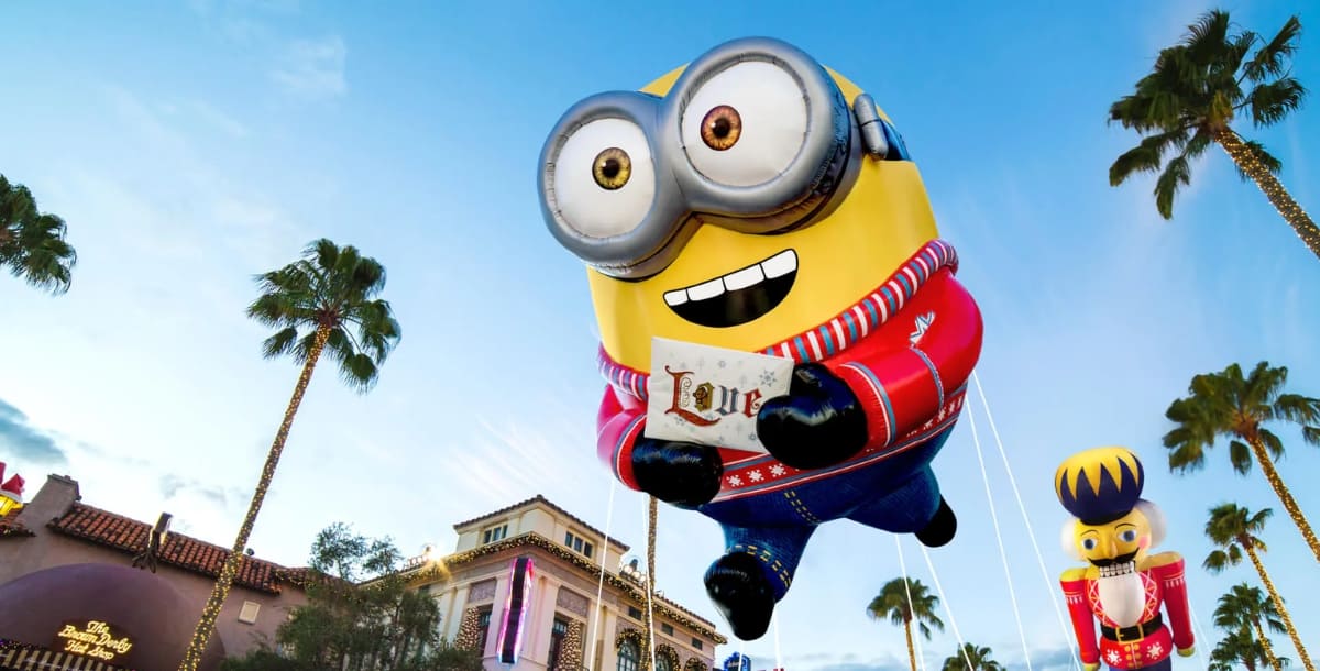 A giant Minion balloon dressed in a festive red sweater floats above Universal Studios, soaring past palm trees and park buildings under a bright blue sky, embodying the playful energy of the beloved Minion popcorn buckets.