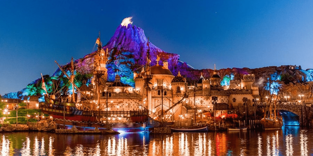 A vibrant, nighttime view of a Disney-inspired theme park featuring a glowing volcano centerpiece, majestic ships docked along the water, and dazzlingly illuminated waterfront buildings—capturing the immersive magic and imaginative atmosphere fans dream of for a potential Disney Fifth Gate.