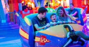 A young family of three enjoys a colorful amusement park ride shaped like a spaceship at Walt Disney World. The child, sitting in the middle, laughs joyfully as the parents, seated on either side, smile and hold onto the ride. Bright, festive lights illuminate the background of this magical Disney attraction.