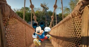 Mickey Mouse stands on a rope bridge surrounded by lush trees and a whimsical wooden structure, welcoming guests to the excitement of runDisney and the splashy adventures awaiting with Water Park Tickets at Disney World.