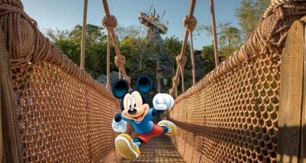 Mickey Mouse stands on a rope bridge surrounded by lush trees and a whimsical wooden structure, welcoming guests to the excitement of runDisney and the splashy adventures awaiting with Water Park Tickets at Disney World.