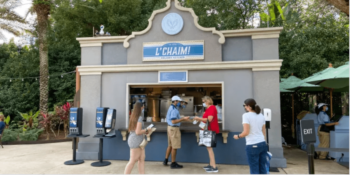 Guests line up to sample holiday treats at the “L’Chaim!” outdoor kiosk, nestled among lush greenery and festive Hanukkah decor, right in the center of Walt Disney World.