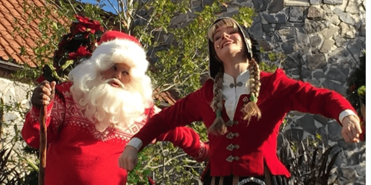 Santa and Mrs. Claus, dressed in their iconic festive red attire, smile for guests outside a picturesque stone building amid lush trees at EPCOT, evoking the magical atmosphere of EPCOT’s holiday storytellers.