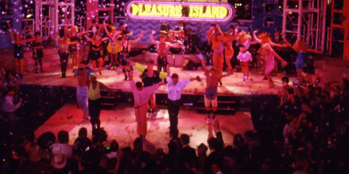 Under the vibrant "Pleasure Island" sign, guests join high-energy dancing on stage as colorful confetti showers down, while a lively crowd cheers and celebrates the entertainment experience.