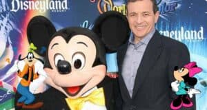 Mickey Mouse greets a guest at a Disneyland Resort event, surrounded by playful Goofy and Minnie Mouse artwork, with Disney CEO Bob Iger in attendance.